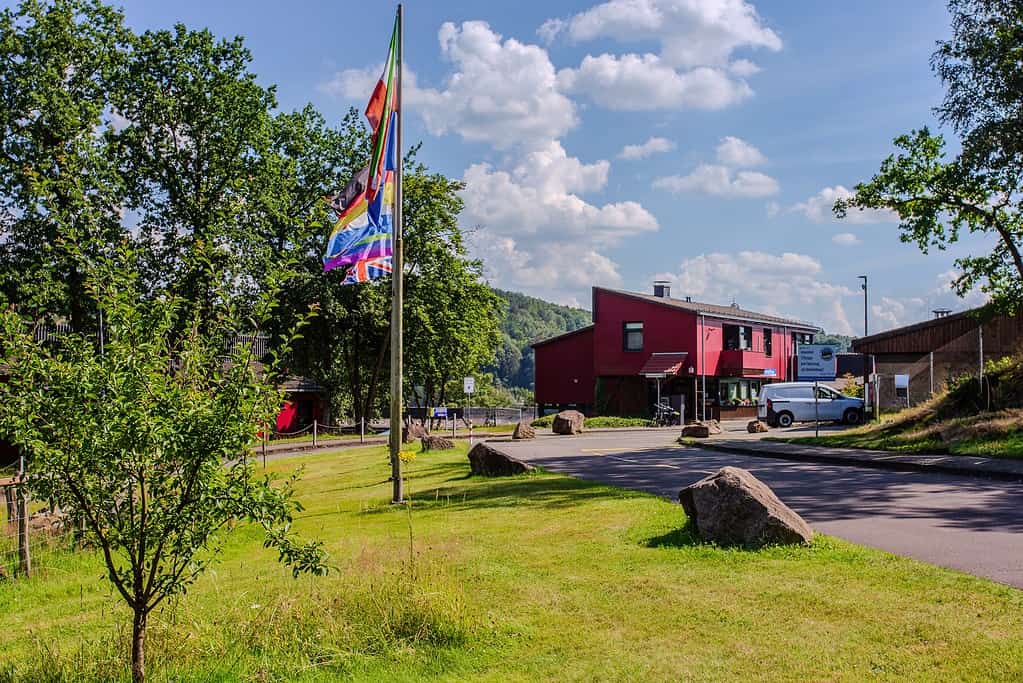 Geschäftshaus am Biggesee mit Flaggen, grüner Rasen, Bäumen, Parkplätzen und Hügel im Hintergrund, bei schönem Wetter.
