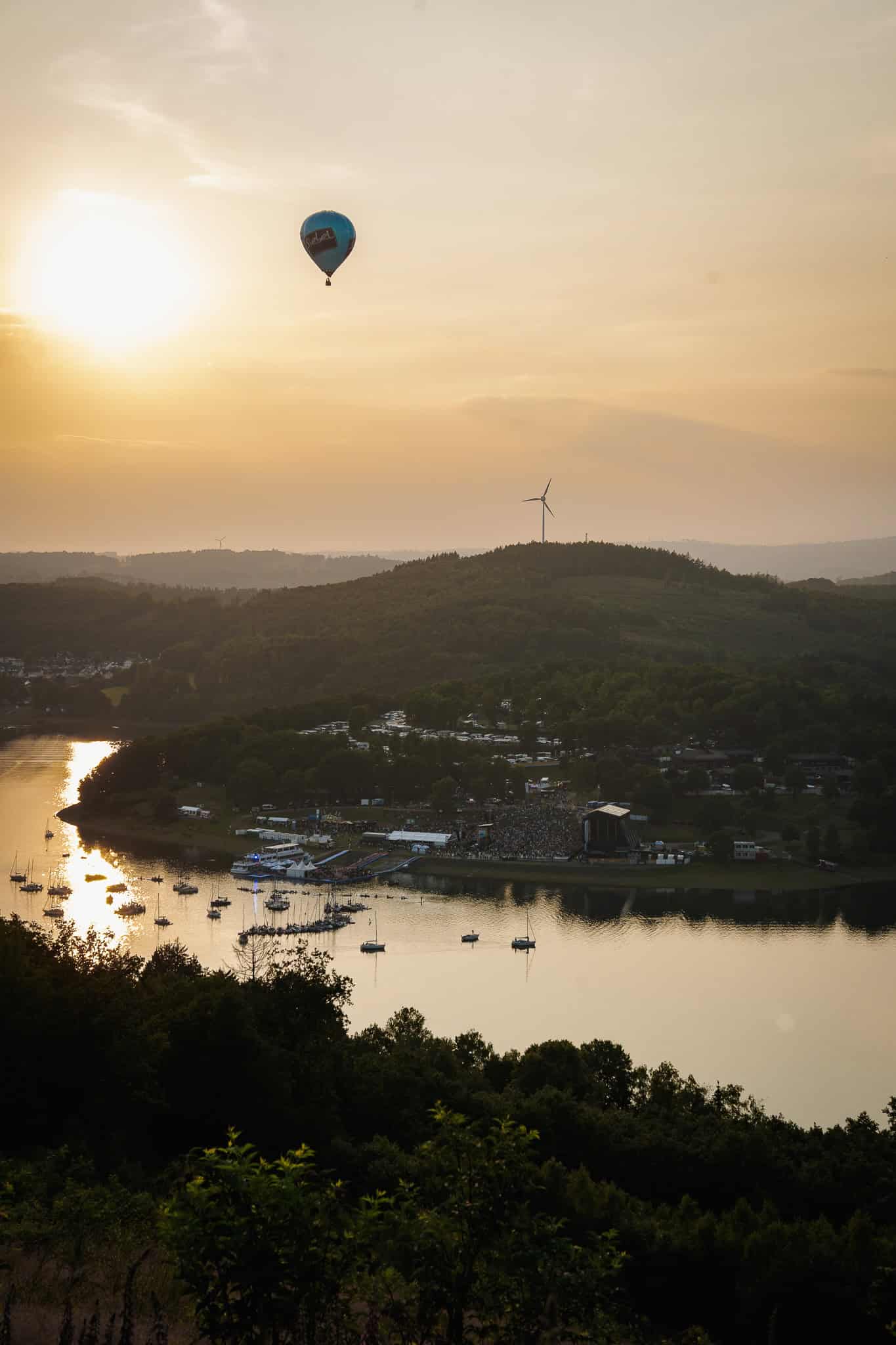 Hochwertige Luftaufnahme des Biggesee bei Sonnenuntergang mit Ballonfahrt während des Biggesee Open Air 2026, umgeben von grünem Landschaft, Wasser und Windkraftanlagen.